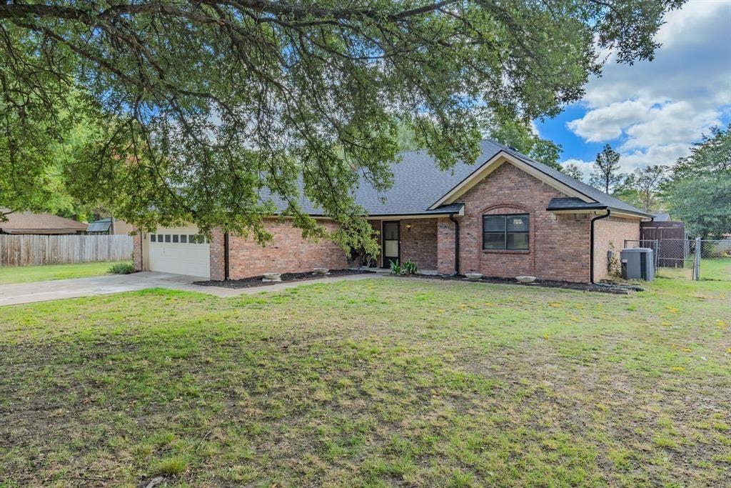 1307 Edna Street, Commerce, TX 75428 Brick exterior featuring a multi-gabled roofline, an attached two-car garage, and a spacious front lawn with mature canopy trees