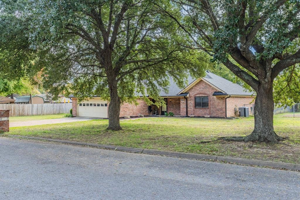 1307 Edna Street, Commerce, TX 75428 Single-story brick exterior featuring a multi-gabled roofline, an attached two-car garage, and mature shade trees
