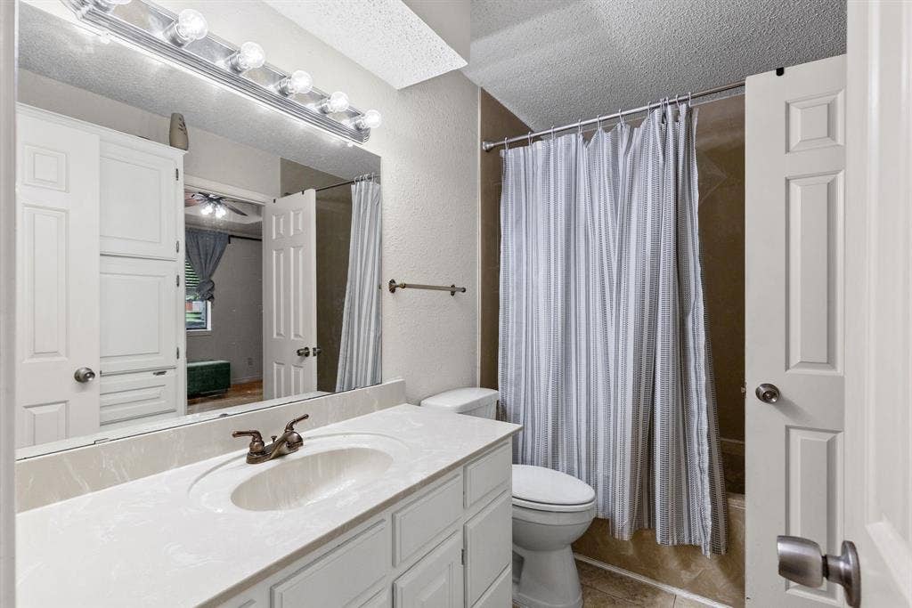 1307 Edna Street, Commerce, TX 75428 Bathroom featuring a stone-composite countertop with an integrated oval basin, white shaker-style cabinetry, wall-mounted multi-bulb light fixture, and a tile-surround shower enclosure