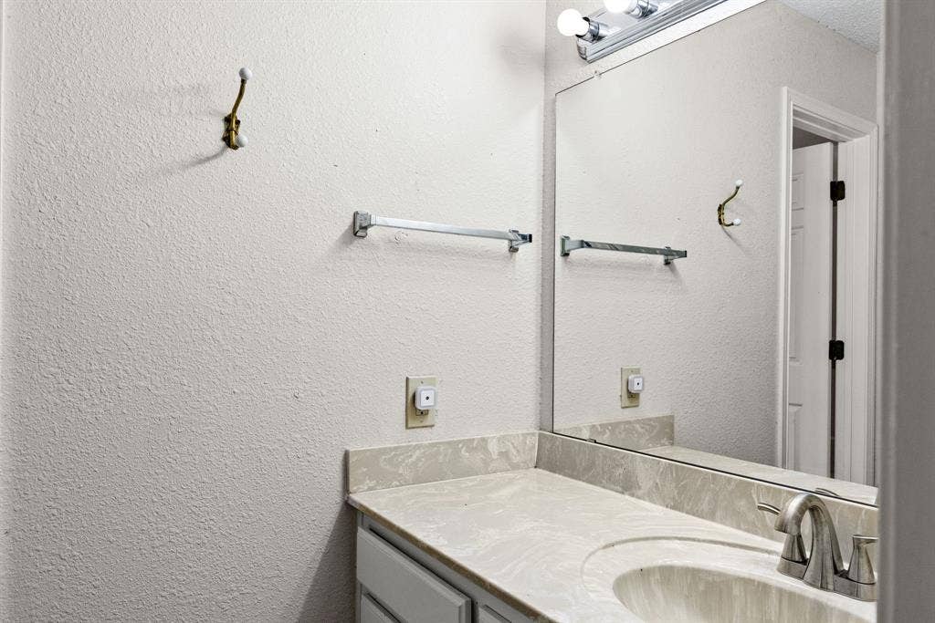 1307 Edna Street, Commerce, TX 75428 Vanity featuring a stone-patterned countertop with an integrated basin, a polished metal faucet, a large frameless mirror, and a multi-bulb light fixture