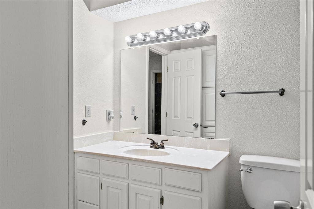 1307 Edna Street, Commerce, TX 75428 Bathroom vanity featuring white cabinetry, a solid-surface countertop with an integrated basin, a large rectangular mirror, and a multi-bulb vanity light fixture
