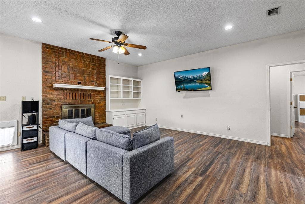 1307 Edna Street, Commerce, TX 75428 Living area featuring wood-finish flooring, floor-to-ceiling brick fireplace with wood mantel, built-in shelving, and ceiling fan with integrated lighting