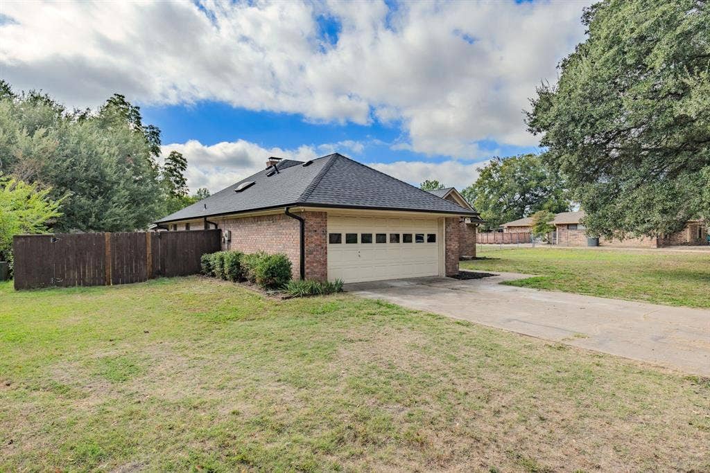 1307 Edna Street, Commerce, TX 75428 Brick exterior with a multi-paneled garage door, asphalt shingle roof, skylights, and a wood-slat privacy fence