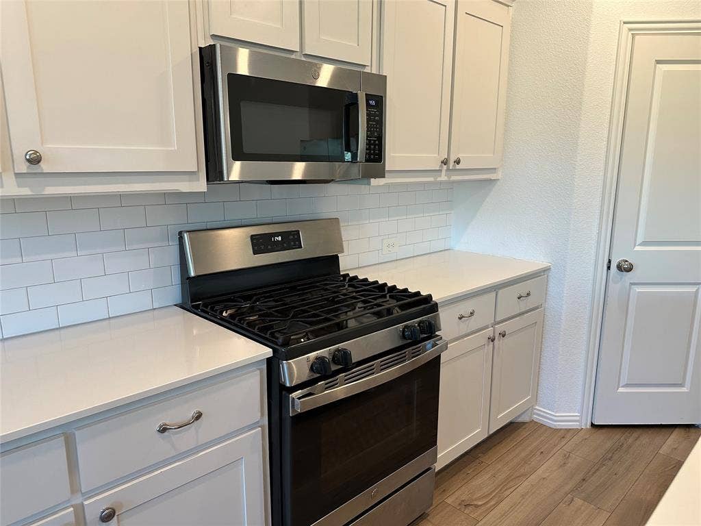 Kitchen featuring white shaker-style cabinetry, stainless steel gas range and microwave, white subway tile backsplash, solid-surface countertops, and wood-finish flooring