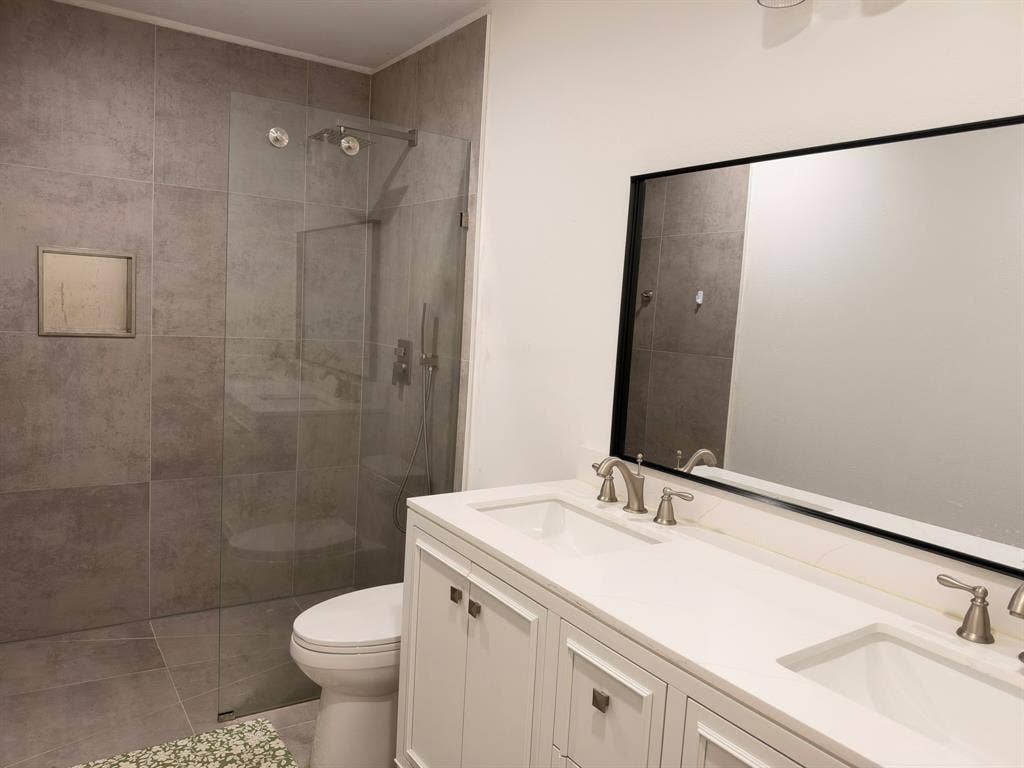 Dual-sink vanity with white cabinetry and countertop, large rectangular mirror with black frame, walk-in shower with floor-to-ceiling tile, and integrated wall niche