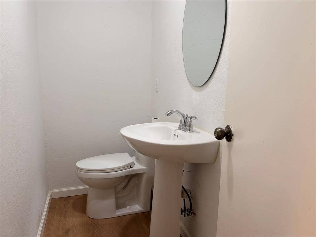 White pedestal sink with chrome faucet, porcelain toilet, oval wall-mounted mirror, and wood-finish flooring