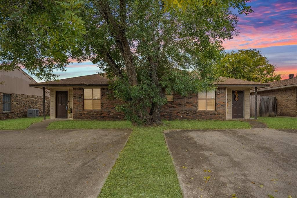 Single-story brick exterior featuring a low-slope roof, dual entry doors, paved surface parking, and mature trees