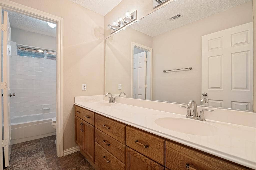 1909 Blackfoot Trail, Mesquite, TX 75149 Dual-sink vanity with wood-finish cabinetry, solid surface countertop, large wall-mounted mirror, and brushed-finish faucets, featuring a separate water closet area with a tub-shower combination and tiled flooring