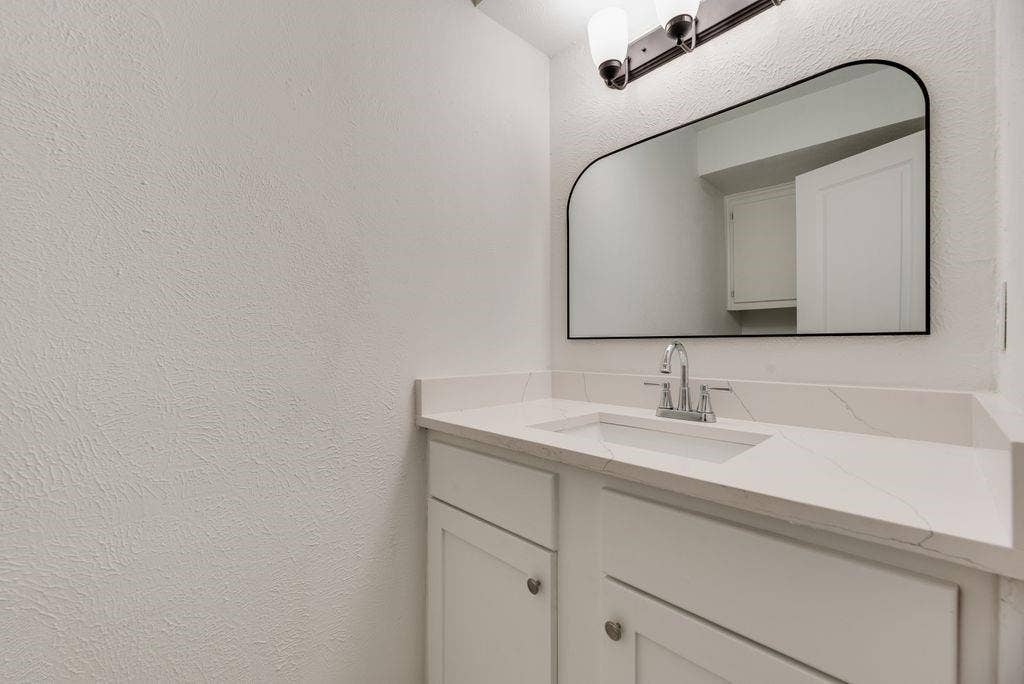 5614 University Hills Boulevard, Dallas, TX 75241 Vanity featuring white cabinetry, quartz countertop, integrated rectangular sink, chrome hardware, and an arched-frame wall mirror