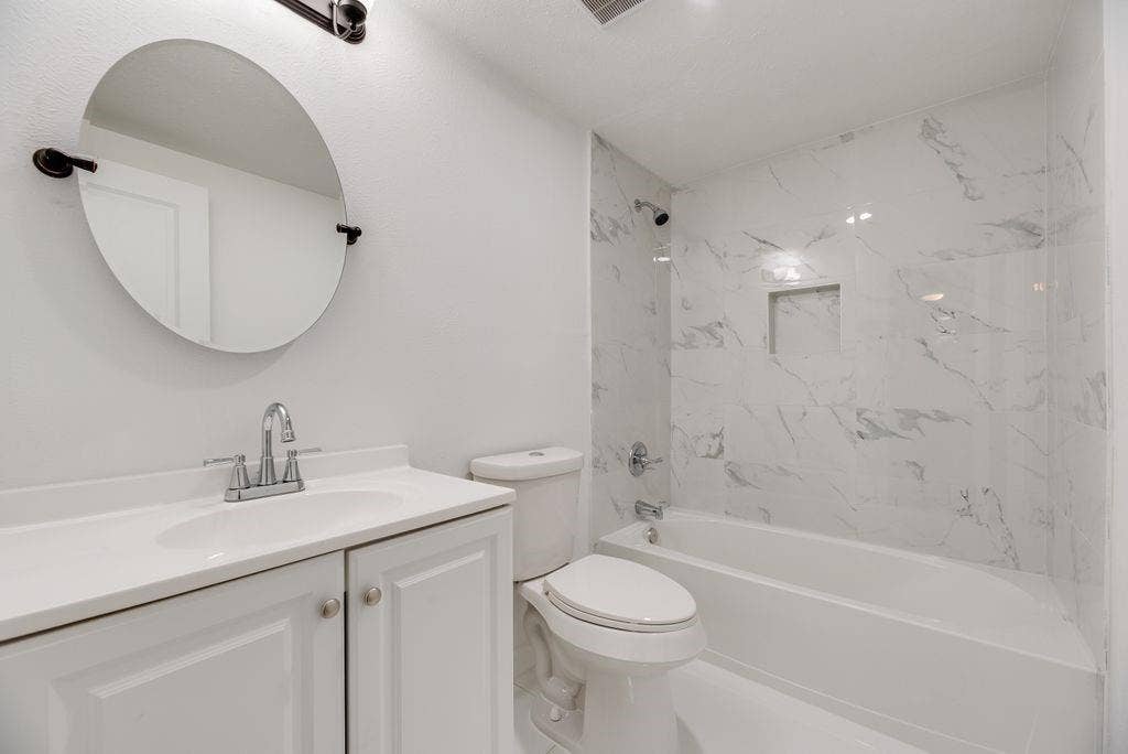 5614 University Hills Boulevard, Dallas, TX 75241 Bathroom featuring a white vanity cabinet with an integrated sink, wall-mounted oval mirror, and a shower-tub combination with white tile surrounds and a built-in recessed niche