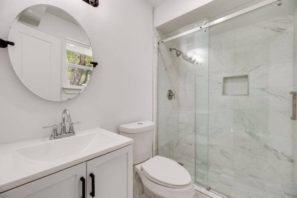 5614 University Hills Boulevard, Dallas, TX 75241 Bathroom featuring a white vanity cabinet, solid-surface countertop, round wall-mounted mirror, and a walk-in shower with stone-style tile surround and a sliding glass door