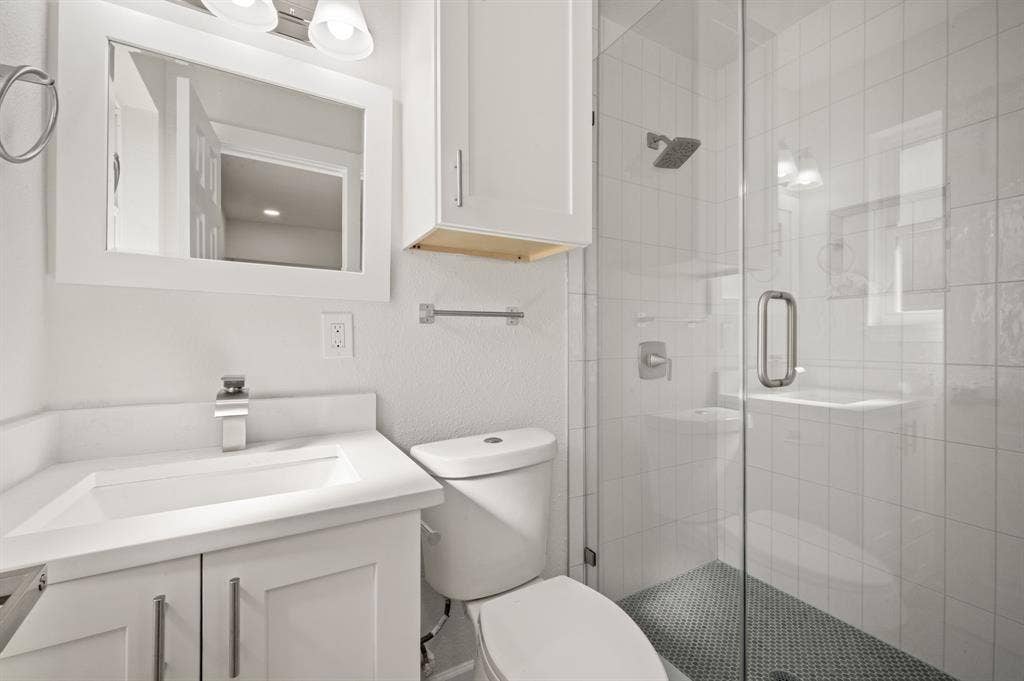 White vanity with a rectangular sink, wall-mounted mirror, white cabinetry, a glass-enclosed shower with tiled surround, and mosaic tile shower flooring