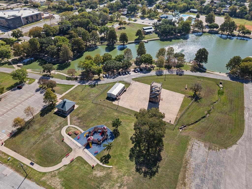 Aerial view of community park grounds featuring a concrete-surfaced area, a dedicated playground structure with cushioned surfacing, a multi-vehicle parking lot, and bordering natural water features