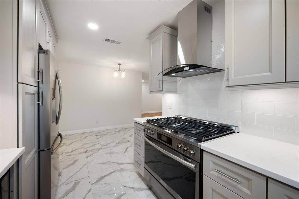 Kitchen featuring light-toned shaker cabinetry, stainless steel gas range with overhead vent hood, white subway tile backsplash, solid surface countertops, and stone-finish flooring