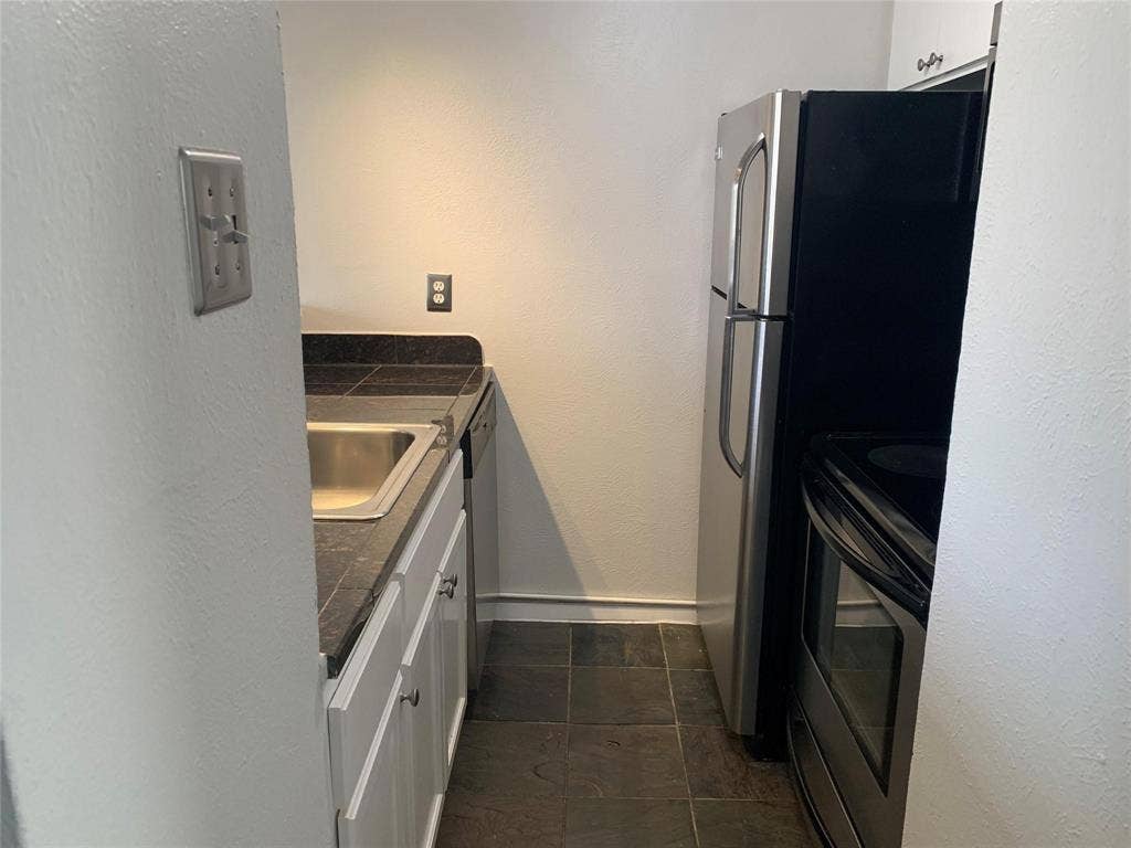 Kitchen featuring white cabinetry, dark countertops, a stainless steel sink, and stainless steel appliances including a refrigerator and oven, complemented by stone-finish flooring