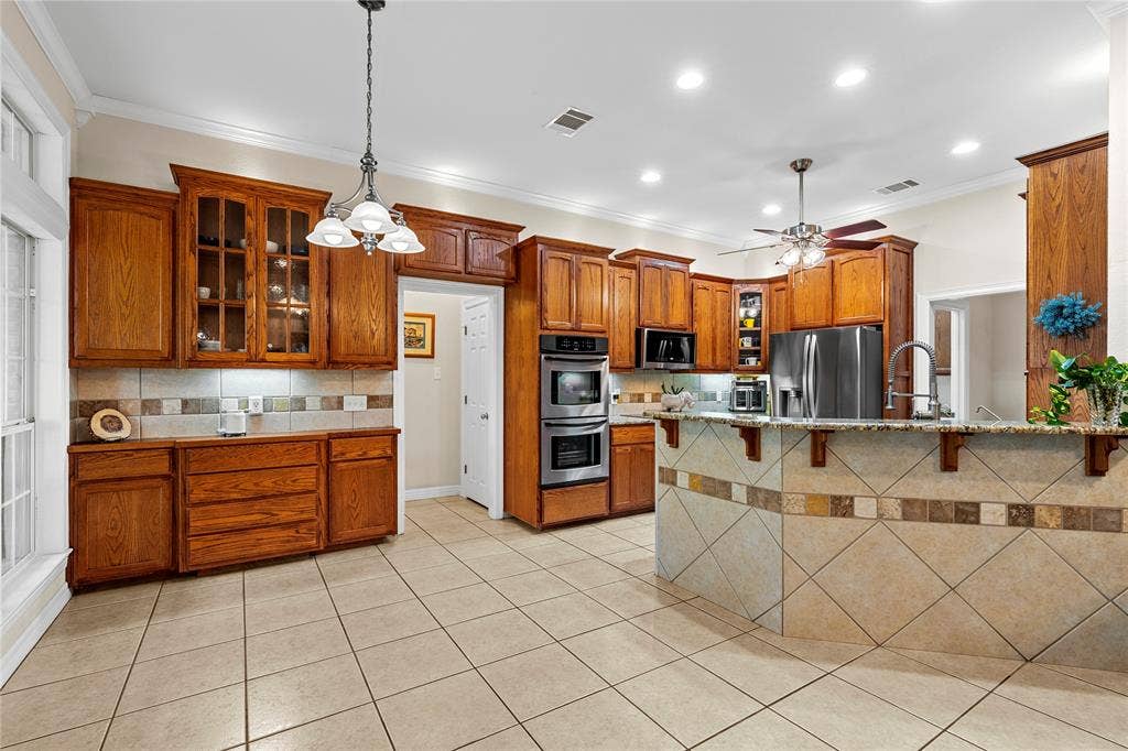Kitchen featuring stained wood cabinetry, stone-tiled backsplash, built-in double wall oven, peninsula with stone-tiled base, and ceramic tile flooring