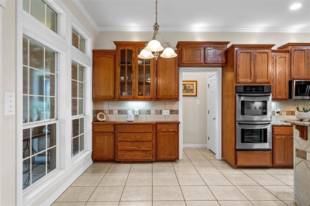 Kitchen featuring wood cabinetry with glass-panel upper doors, stainless steel double wall ovens, tile flooring, tile backsplash, and a multi-light pendant fixture