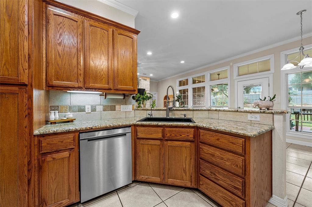 Kitchen featuring stained wood cabinetry, stone-surface countertops, multi-tonal tile backsplash, stainless steel dishwasher, and a dark-finish drop-in sink with a high-arch faucet