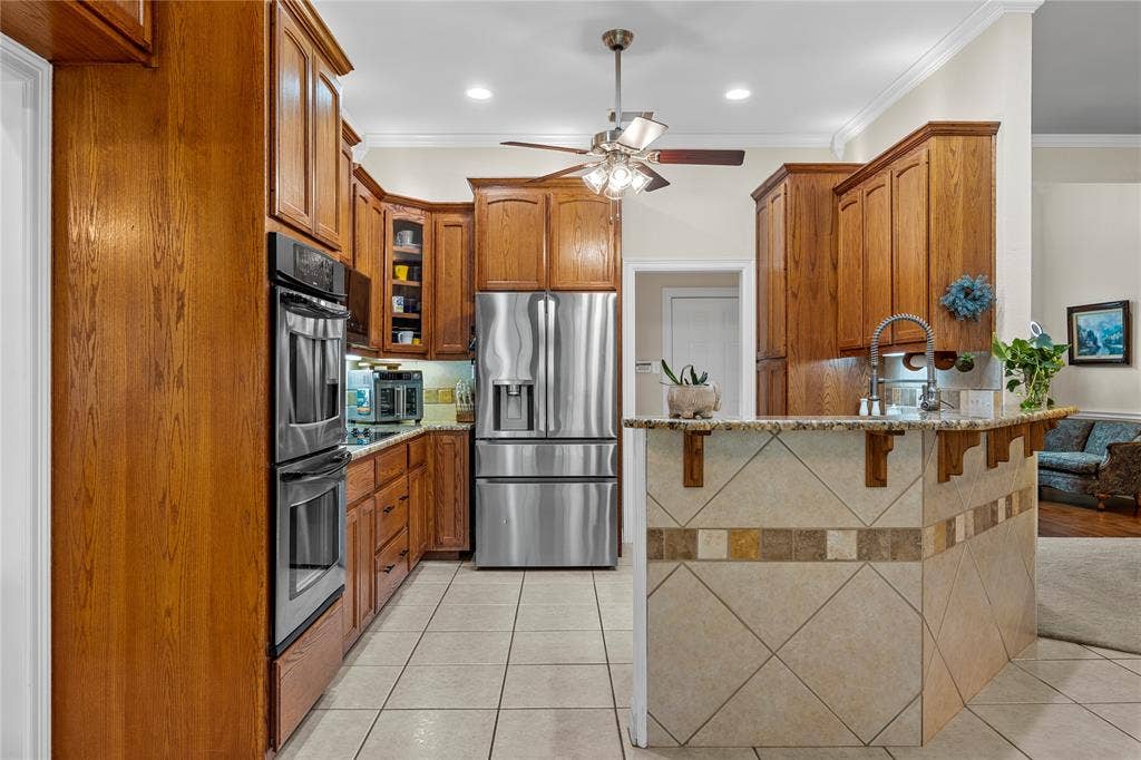 Kitchen featuring wood cabinetry, stainless steel double wall ovens and refrigerator, stone-surfaced island with tile facing, and tile flooring