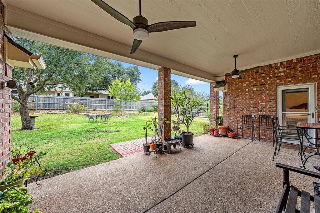 Covered patio featuring an exposed aggregate concrete floor, red brick exterior wall, and dual overhead ceiling fans, extending to a fenced backyard