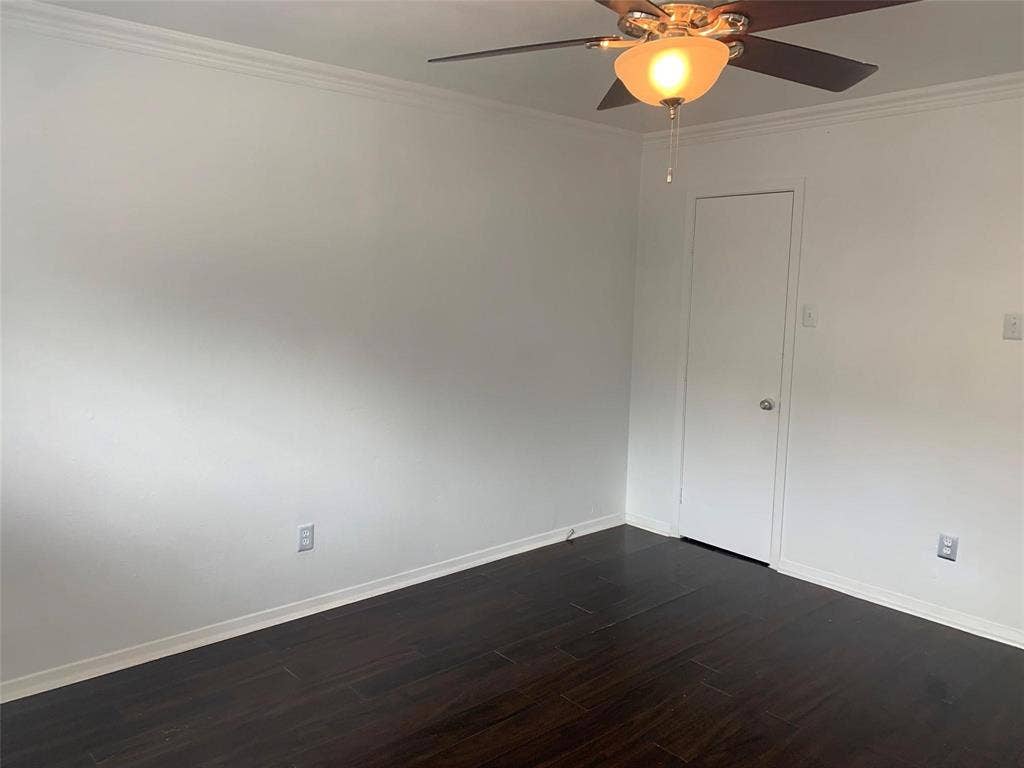 Interior room featuring dark wood-finish flooring, neutral wall paint, crown molding, and an attached ceiling fan with a glass light fixture
