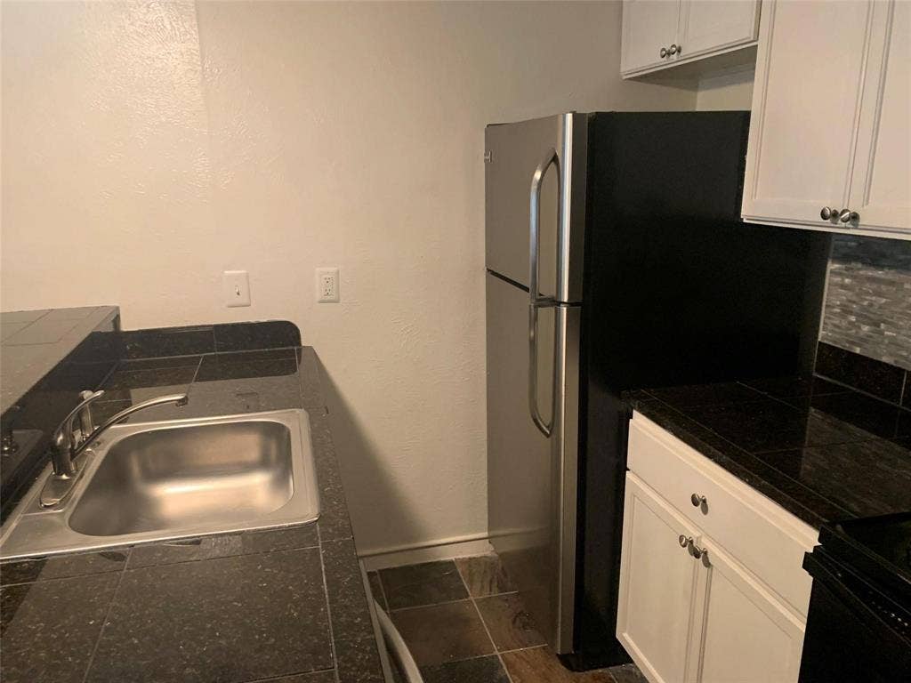 Kitchen area featuring dark tile countertops, stainless steel undermount sink, white cabinetry, and stainless steel refrigerator