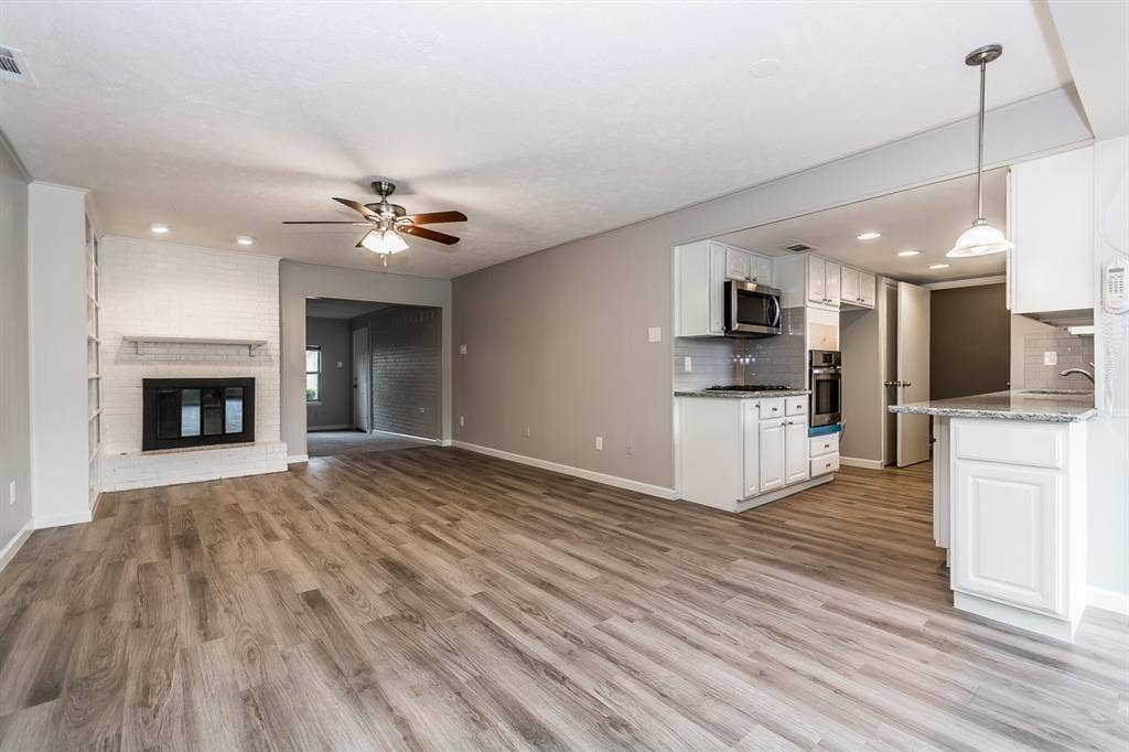 Open floor plan featuring wood-finish flooring, a white brick fireplace, recessed lighting, and a kitchen equipped with white cabinetry, stone-finish countertops, and built-in stainless steel appliances