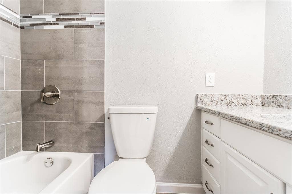 Bathroom featuring a vanity with a light-toned stone countertop, white cabinetry with silver-tone hardware, a bathtub with grey tile surround, and a decorative glass and stone mosaic accent border