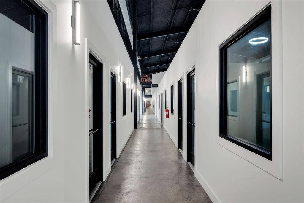 Industrial-style corridor featuring polished concrete flooring, exposed high-ceiling trusses, crisp white walls, floor-to-ceiling glass-paneled doors, and modern wall-mounted linear light fixtures