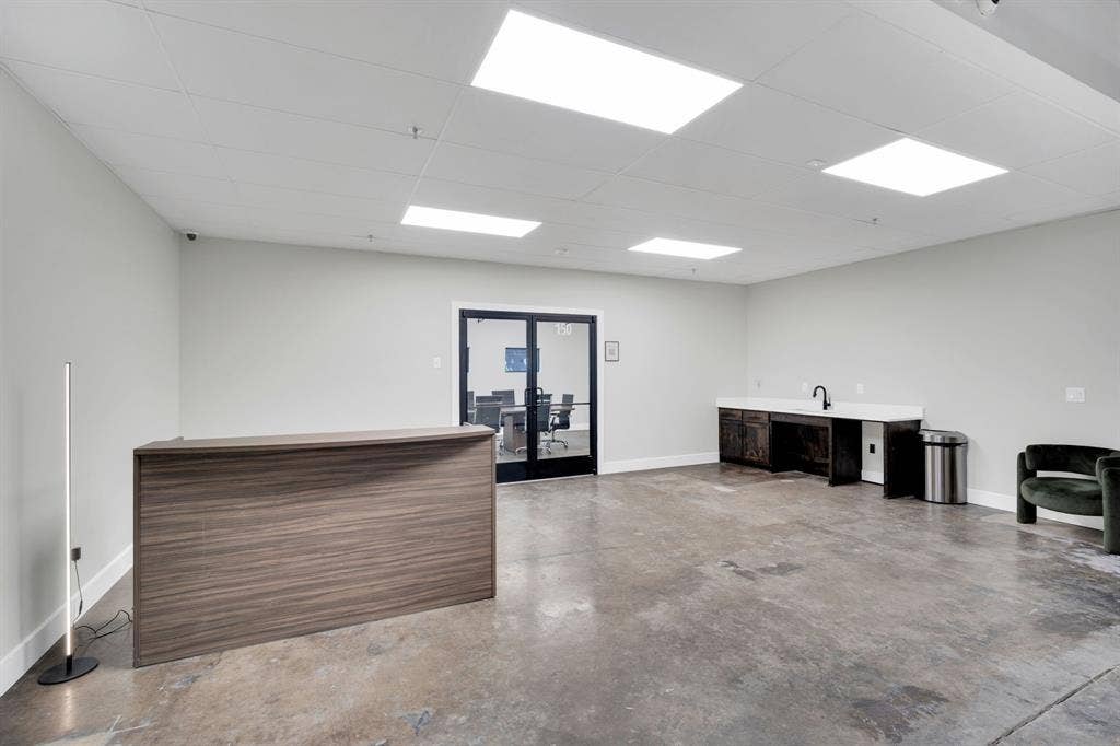 Open-concept interior featuring stained concrete flooring, recessed panel ceiling lighting, a built-in wood-finish reception desk, a kitchenette with stone-surface countertop, and glass-paned double doors