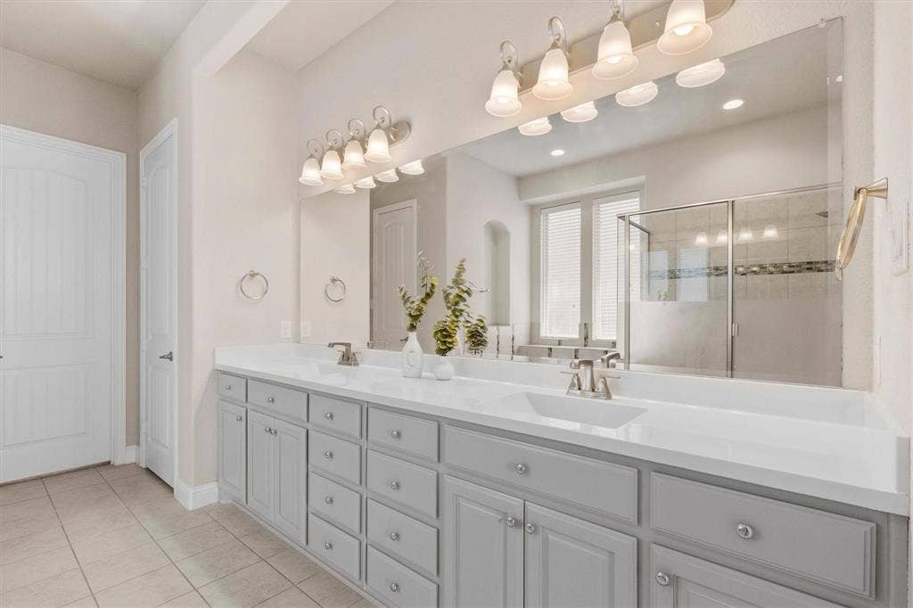 4109 Rainey Street, McKinney, TX 75070 Dual-sink vanity with light-gray cabinetry, expansive countertop, full-width wall mirror, and two multi-bulb overhead light fixtures