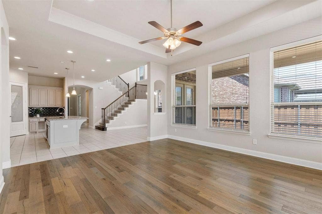 4109 Rainey Street, McKinney, TX 75070 Open-concept interior featuring wood-finish flooring, tray ceilings with a central fan, large windows, a staircase with metal balusters, and a kitchen island with stone countertops
