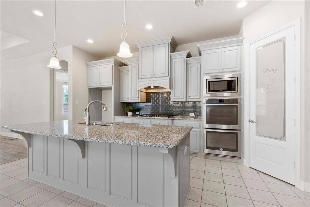 4109 Rainey Street, McKinney, TX 75070 Kitchen featuring light gray cabinetry, expansive stone-topped island, stainless steel double wall ovens, glass-tiled backsplash, and pantry door with frosted glass panel