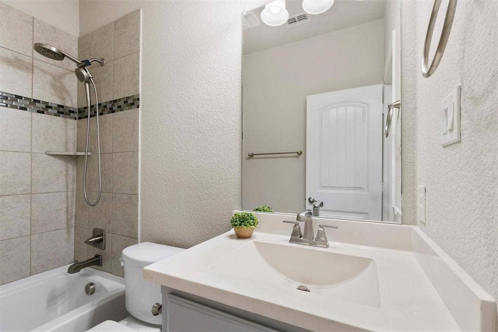 4109 Rainey Street, McKinney, TX 75070 Vanity with a solid-surface countertop and integrated sink, complemented by a handheld shower fixture, tiled tub surround with decorative mosaic accent, and a full-width wall-mounted mirror