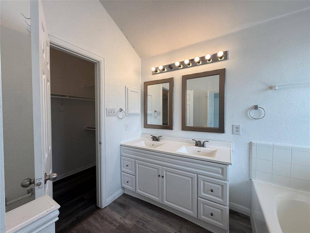 Dual-sink vanity with white cabinetry and solid-surface countertop, twin rectangular mirrors, multi-bulb vanity lighting, and wood-finish flooring
