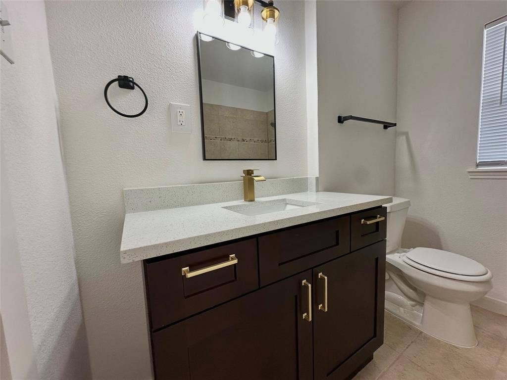 Dark wood cabinetry featuring gold-tone hardware, white stone countertop, rectangular undermount sink, matching gold-tone faucet, and three-light vanity fixture