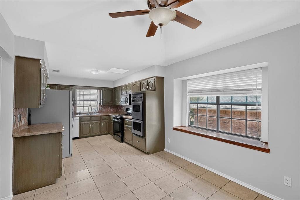 2413 Club Meadow Drive, Garland, TX 75041 Spacious kitchen featuring stainless steel appliances, wood-finish cabinetry, tile flooring, and abundant natural light from large windows with wood-trimmed sills and window seat