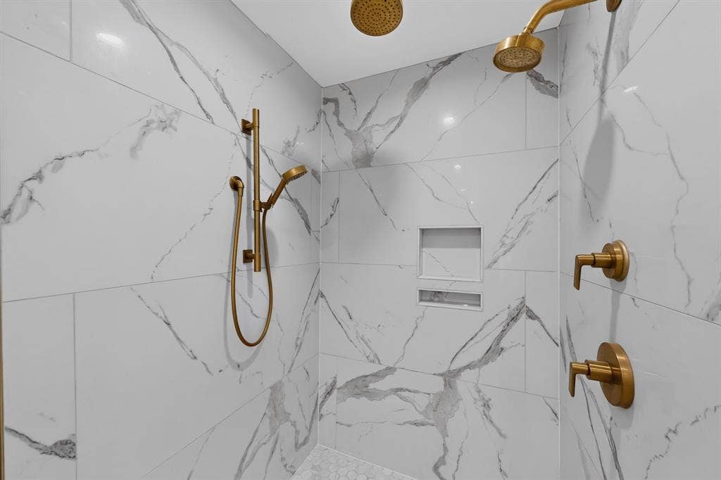 2413 Club Meadow Drive, Garland, TX 75041 White and grey veined tile shower walls, dual shower heads, built-in recessed shelving, gold-finish fixtures, and a handheld shower