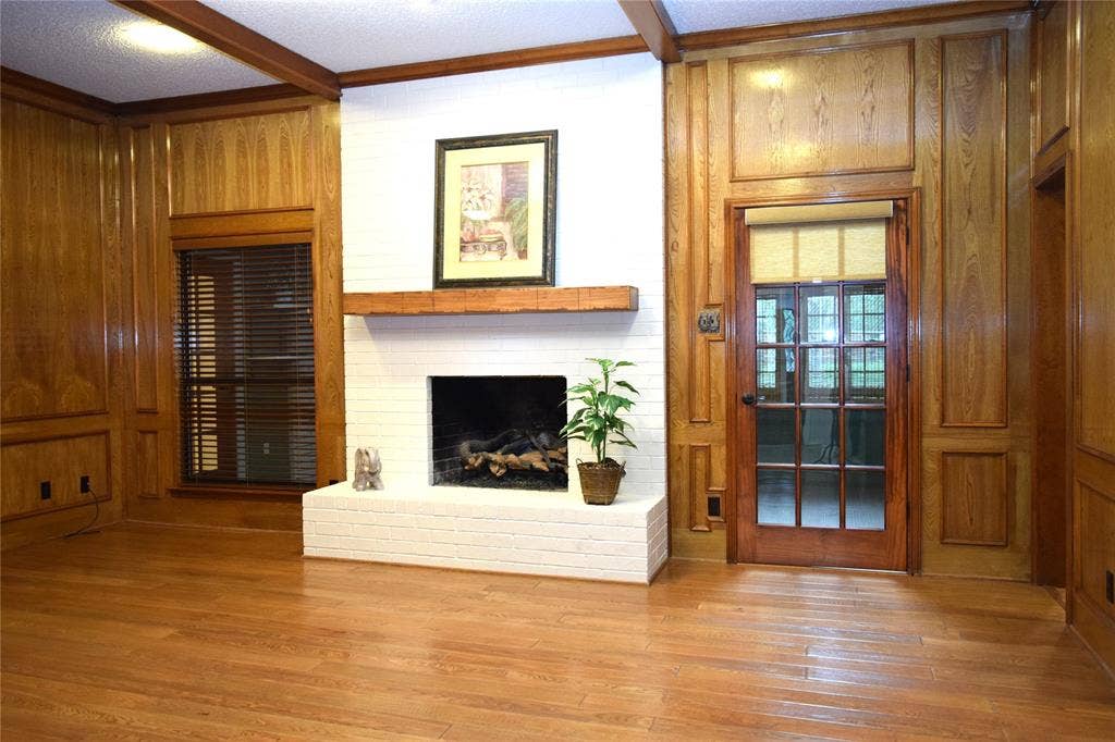 Wood paneling with recessed molding, white-painted brick fireplace, timber beam ceiling accents, wood-finish flooring, and French glass door with wood trim