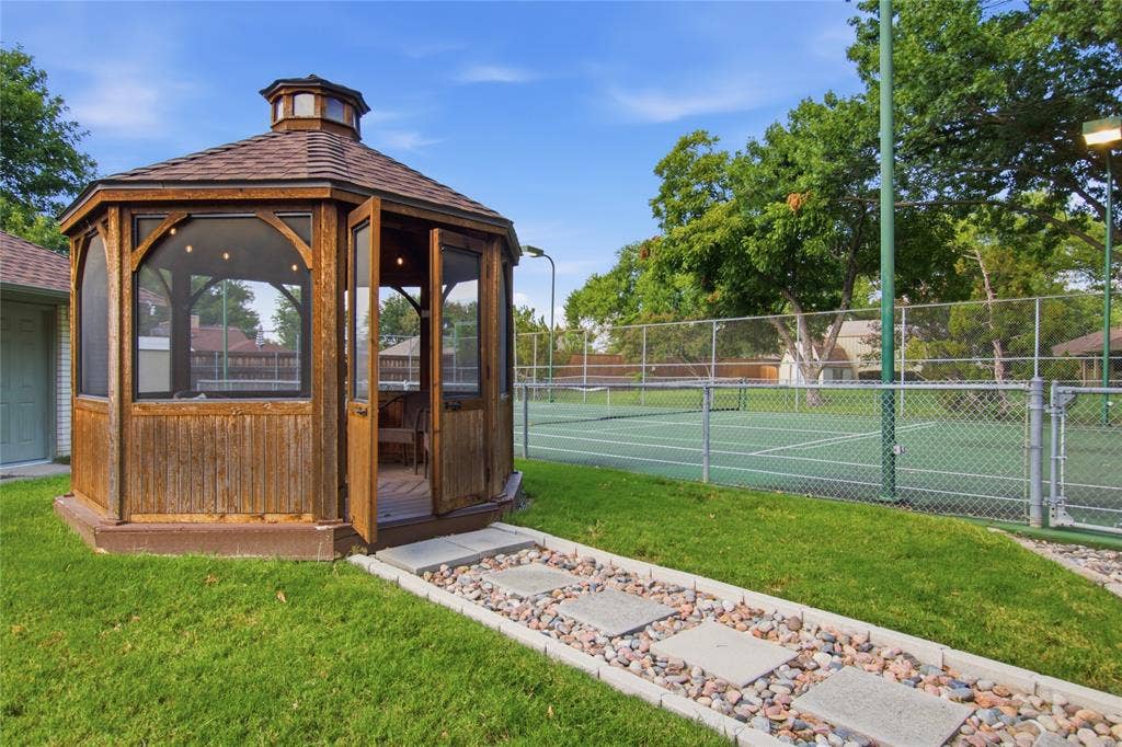 Octagonal wood gazebo with a shingled roof and screened panels, paired with a fenced hard-surface tennis court and a stone paver garden path