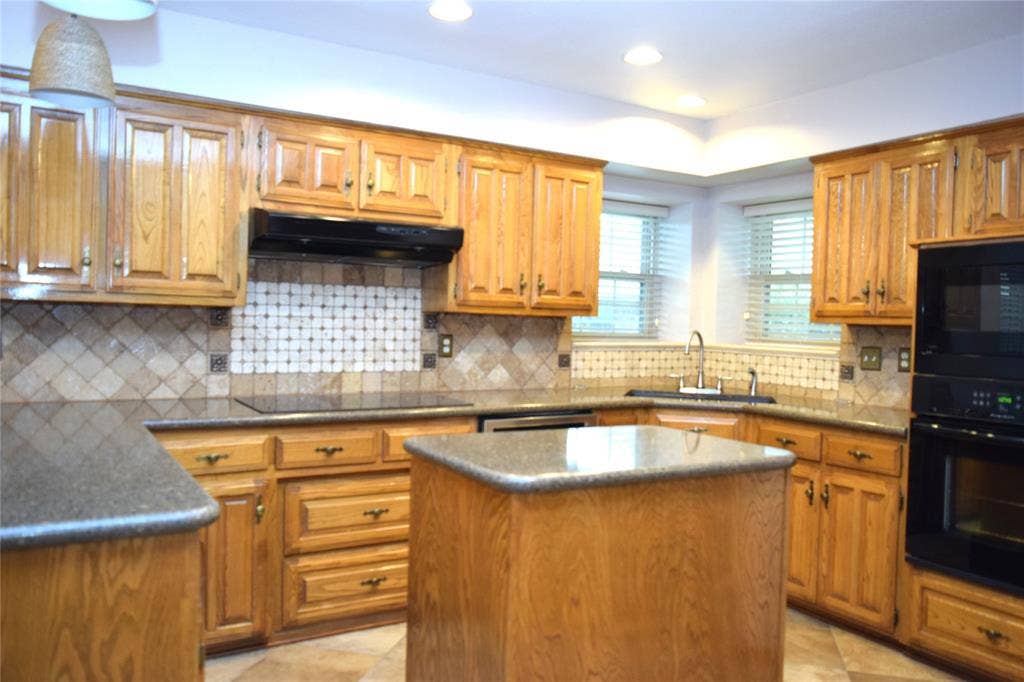 Kitchen featuring wood cabinetry, stone-finish countertops, a central island, tile backsplash with inset accent detailing, and built-in black wall ovens