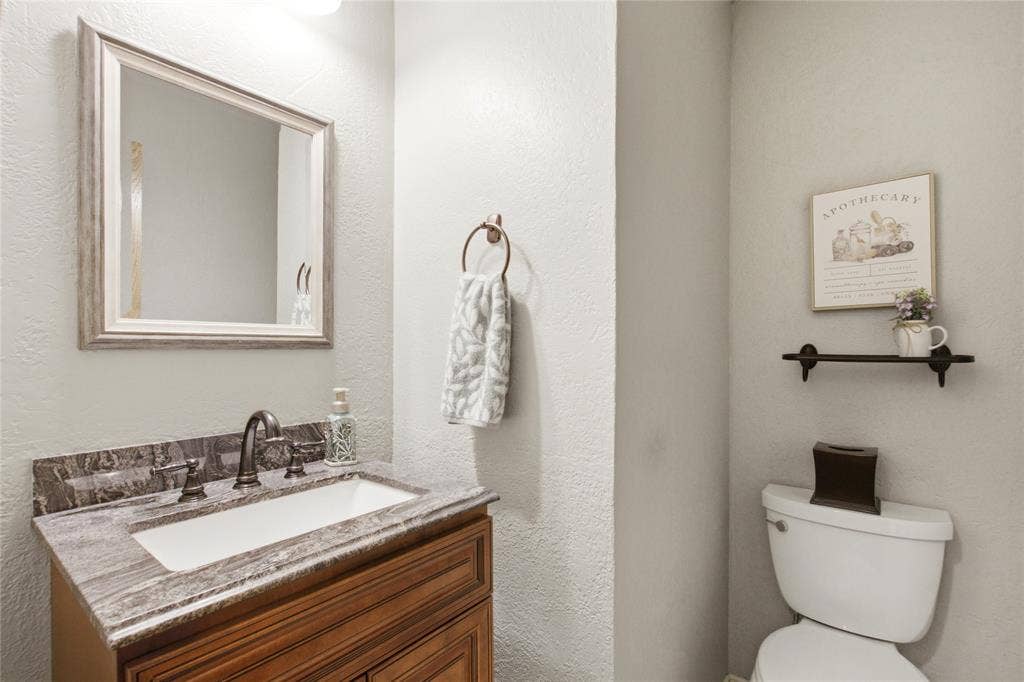Wood-finish vanity cabinet, stone-patterned countertop with an integrated rectangular sink, antique-style bronze faucet, framed wall mirror, and dual-flush toilet