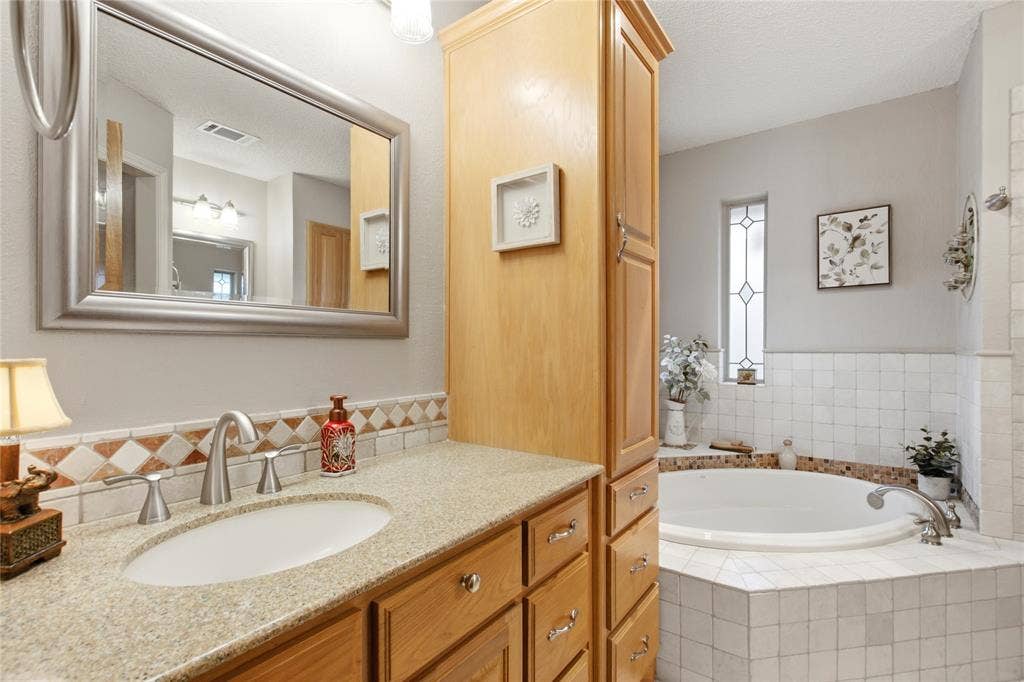 Bathroom featuring a natural wood-finish vanity, stone-composite countertop, full-height storage cabinetry, an inset soaking tub, and decorative tile accents