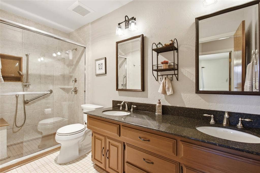 Dual-sink vanity with dark stone countertop, wood cabinetry, glass-enclosed shower, and framed wall mirrors