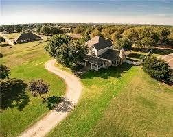 Aerial perspective of a multi-story residence featuring a winding driveway, expansive acreage, mature trees, and a covered porch