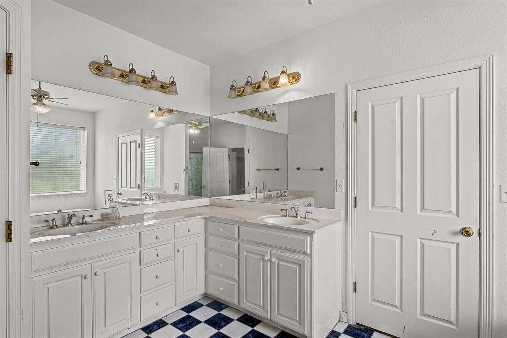 L-shaped vanity featuring white cabinetry, dual undermount sinks, stone countertops, expansive wall-to-wall mirrors, and checkered tile flooring