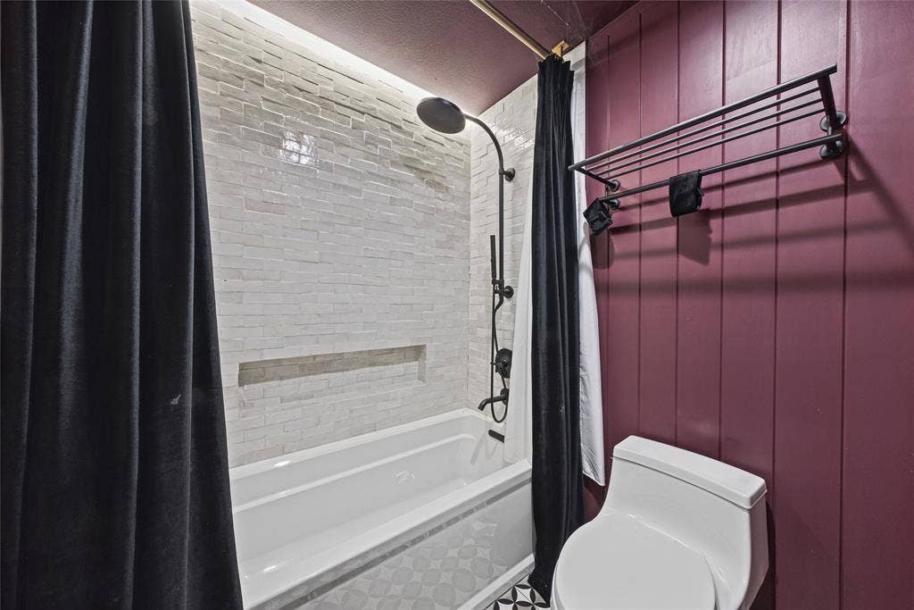 605 Ranier Circle, Garland, TX 75041 Bathtub featuring textured tile surround with recessed niche, matte black rainfall shower system, vertical wood-panel wall, and wall-mounted metal shelving