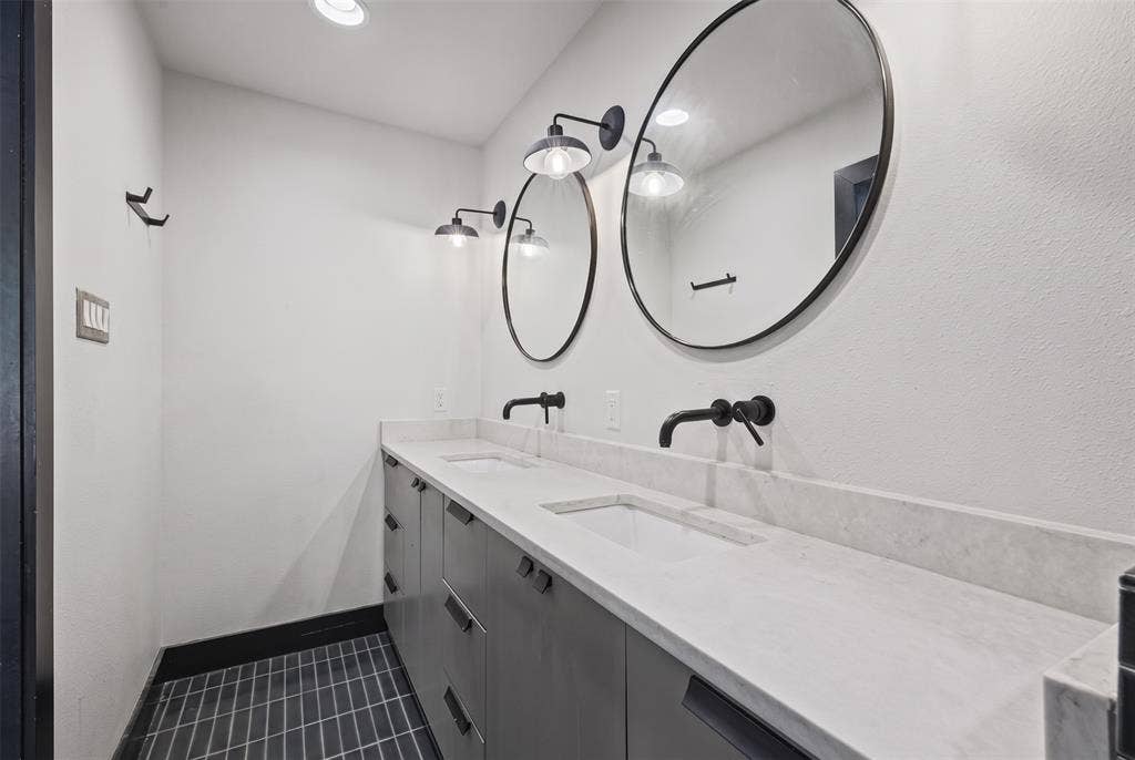 605 Ranier Circle, Garland, TX 75041 Dual-sink vanity featuring charcoal-finished cabinetry, a white stone countertop, wall-mounted matte black faucets, round mirrors, and industrial-style wall sconces