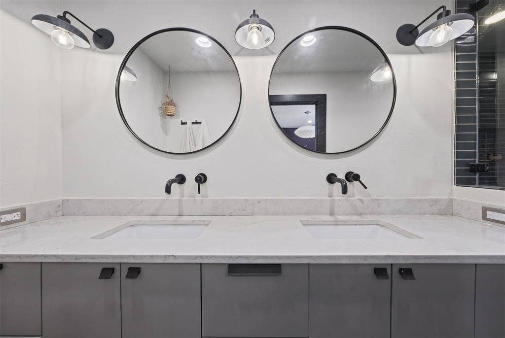 605 Ranier Circle, Garland, TX 75041 Dual-sink vanity featuring gray flat-panel cabinetry, light-toned stone countertops with integrated rectangular basins, matte black wall-mounted faucets, dual round mirrors, and matching industrial-style sconces