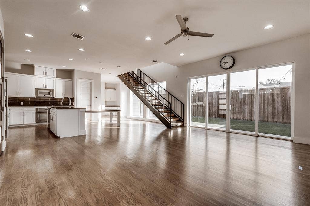 Open-concept interior featuring wood-finish flooring, recessed lighting, a kitchen with white cabinetry and tiled backsplash, a staircase with metal railings, and sliding glass doors